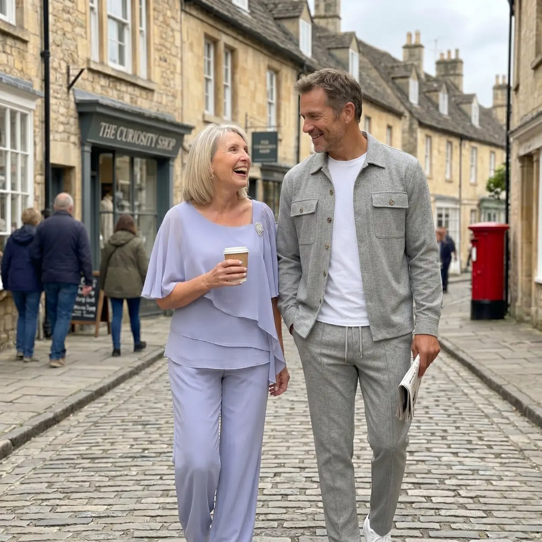 Man and woman walking together on a cobblestone street, holding coffee and a newspaper.